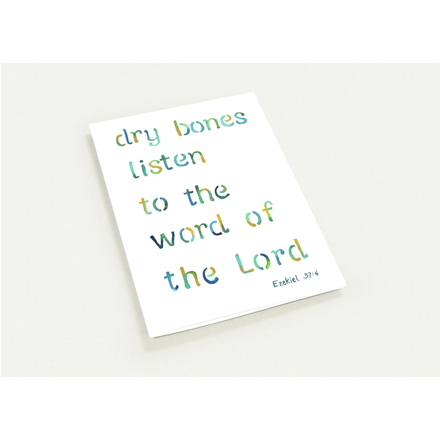 A white rectangular art card with the phrase 'dry bones listen to the word of the Lord' in multicolored hand-lettered text, plus 'Ezekiel 37:4' in small black script at the bottom right, photographed on a light neutral background.