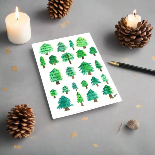 Greeting card with watercolor painted green Christmas trees set on a gray desk beside candles and pinecones.