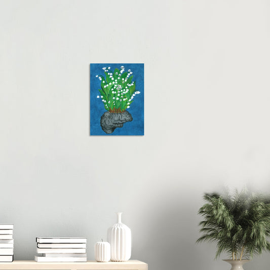 Print of a floral design of lilies coming out of a brain on a blue background, displayed on a wall with stacked books on a desk and green plant beside it