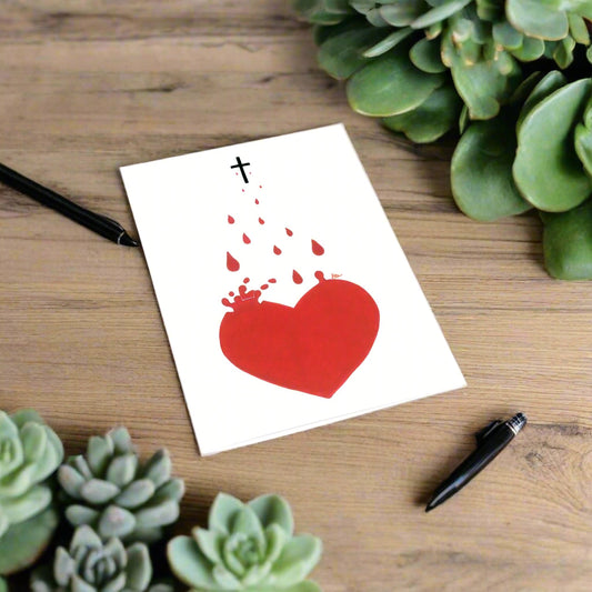 A white rectangular card with a red heart and a small black cross at the top, photographed on a wooden surface with succulents and two black pens around it.