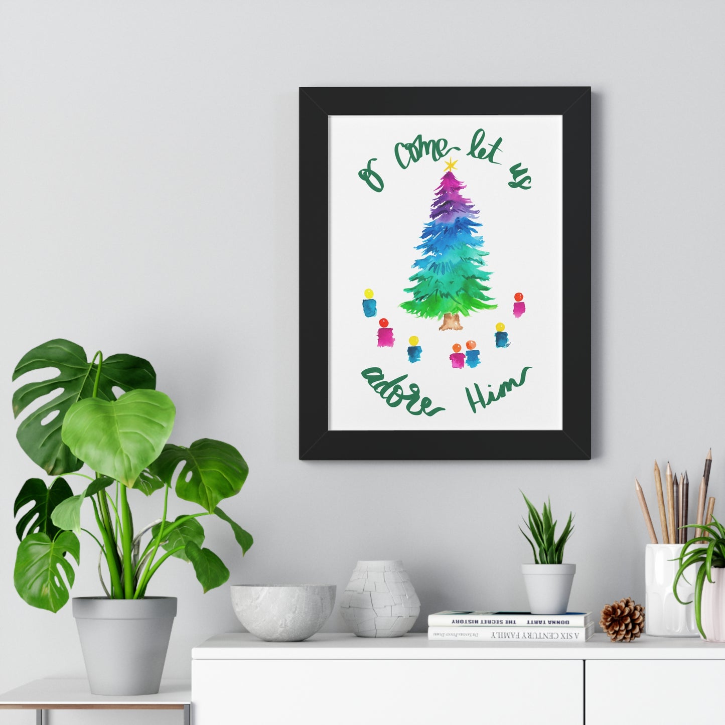 Framed artwork with a colorful Christmas tree and text “O come let us adore Him” on a wall above a white cabinet with decorative items.