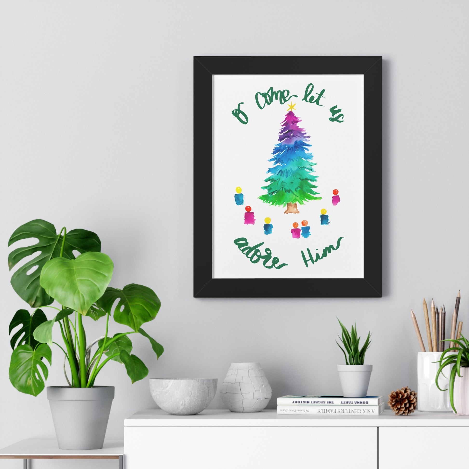 Framed artwork with a colorful Christmas tree and text “O come let us adore Him” on a wall above a white cabinet with decorative items.