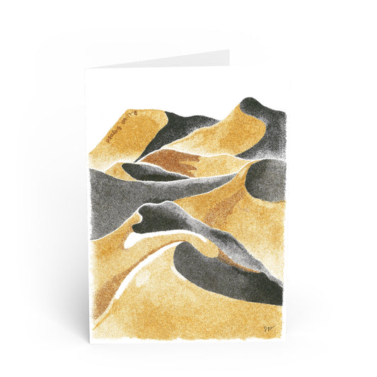 A folded greeting card standing upright on a white background, featuring an abstract desert dune illustration in textured gold, black, and gray tones.