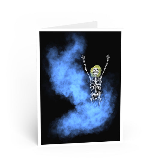 A folded greeting card standing upright on a white background. The card design shows a skeleton with raised arms floating in blue smoke against a black background.
