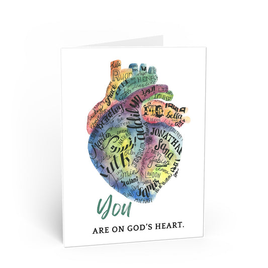 A white greeting card shown standing upright on a white background. The front features a colorful typographic illustration of a heart made up of names, with the words 'You ARE ON GOD'S HEART.