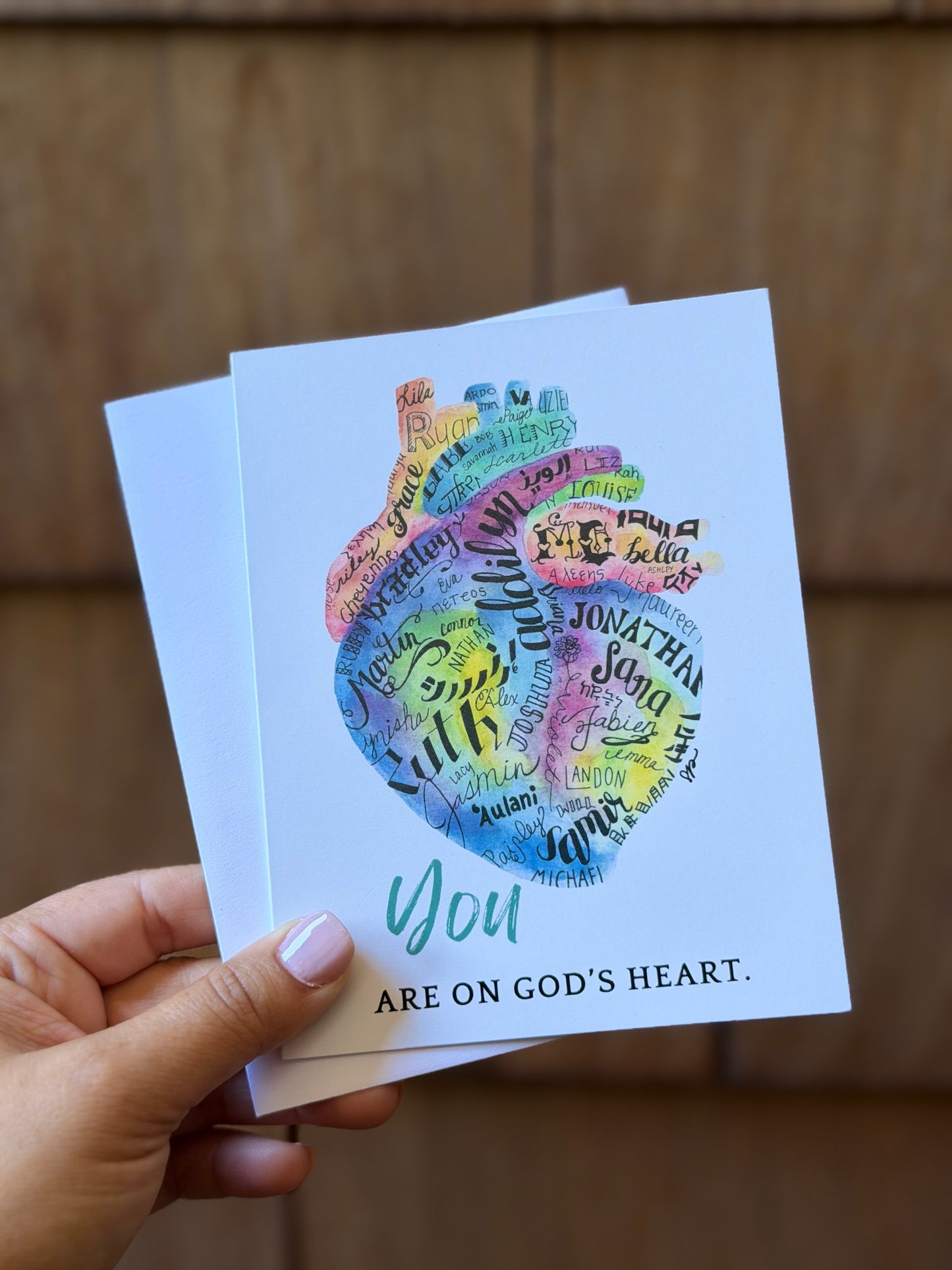 A hand holds two white greeting cards against a blurred brown wooden background. The front card features a colorful heart made of handwritten names and the text 'You ARE ON GOD'S HEART.