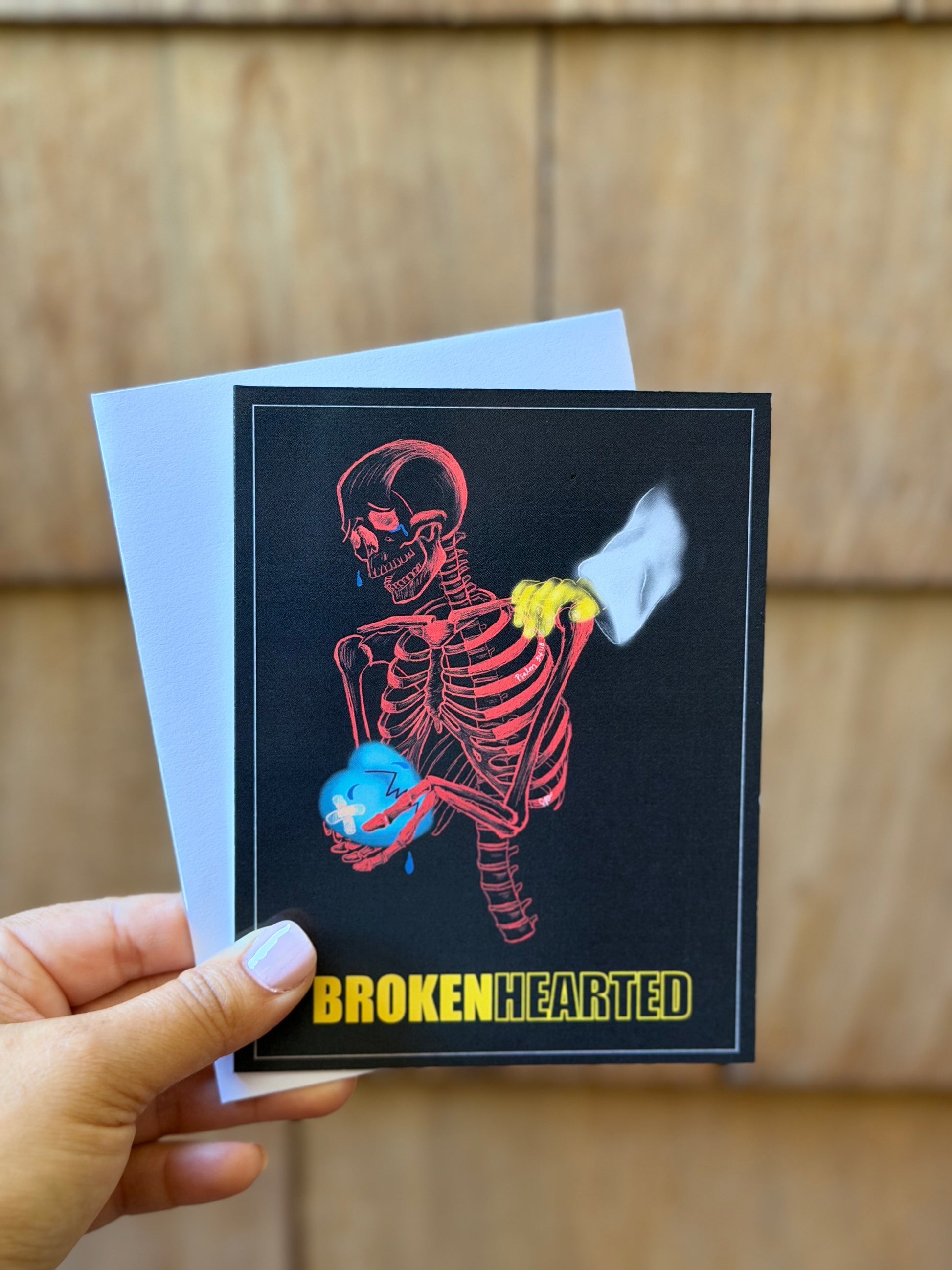 A hand holds a square black greeting card featuring a red skeleton holding a blue heart, with a white broken heart graphic and the text 'BROKEN HEARTED' at the bottom. A white envelope is partially visible behind the card, set against a blurred wood-paneled background.