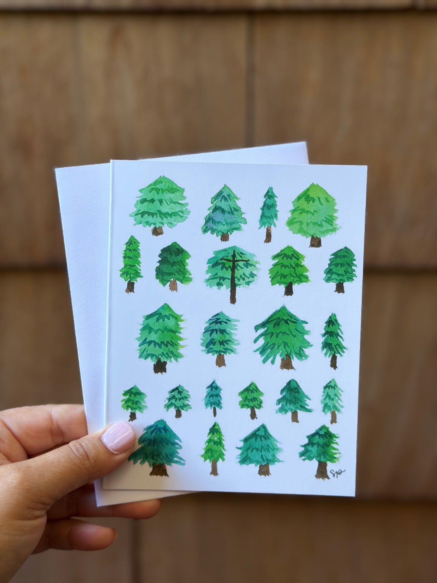 A hand holds a white greeting card featuring watercolor illustrations of various green pine trees, with a matching white envelope behind it, set against a warm brown wooden background.