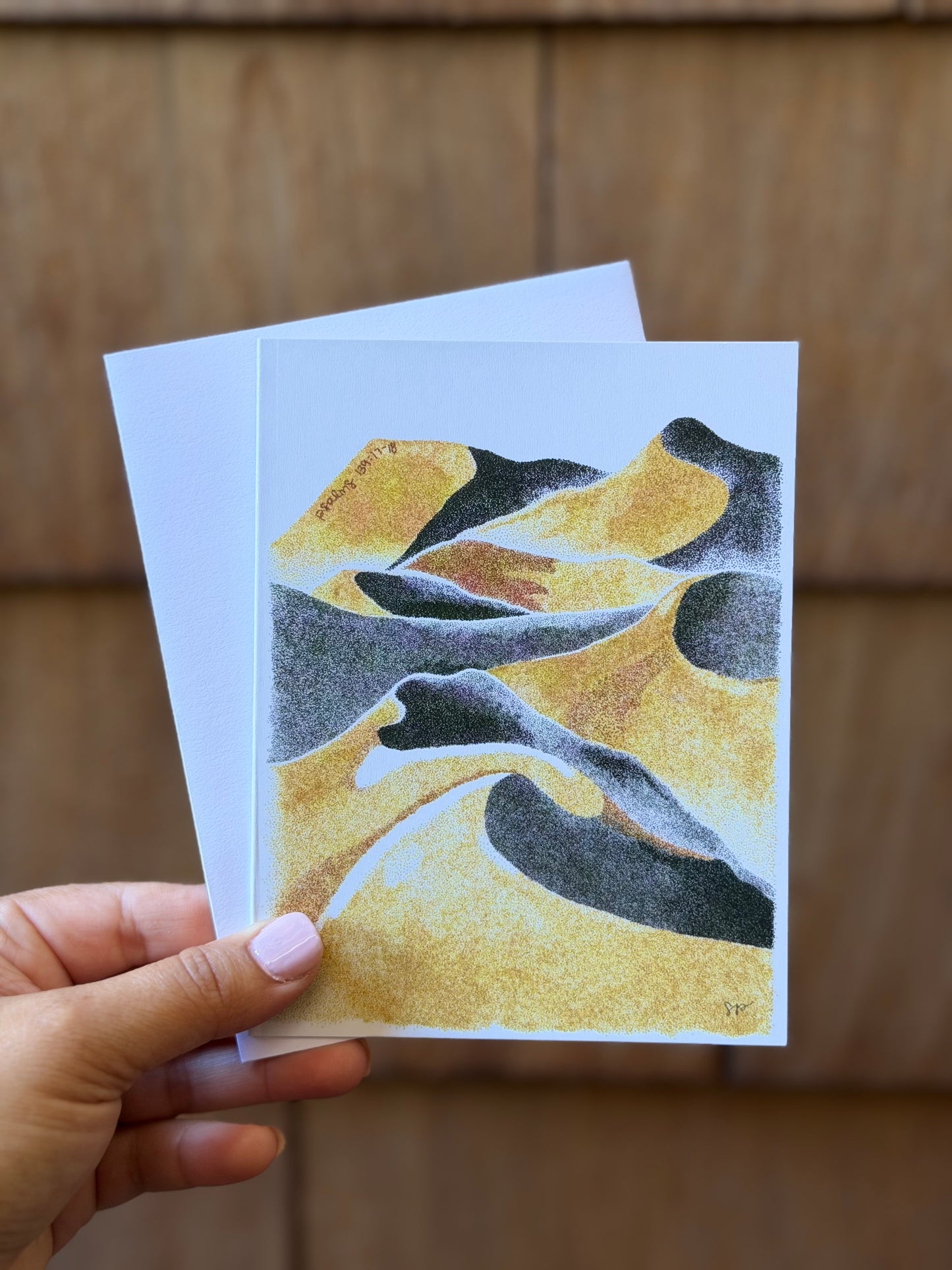 A hand holds a small art print or card featuring an abstract desert dune design in gold, black, and gray, with a white backing card behind it against a blurred brown wooden background.