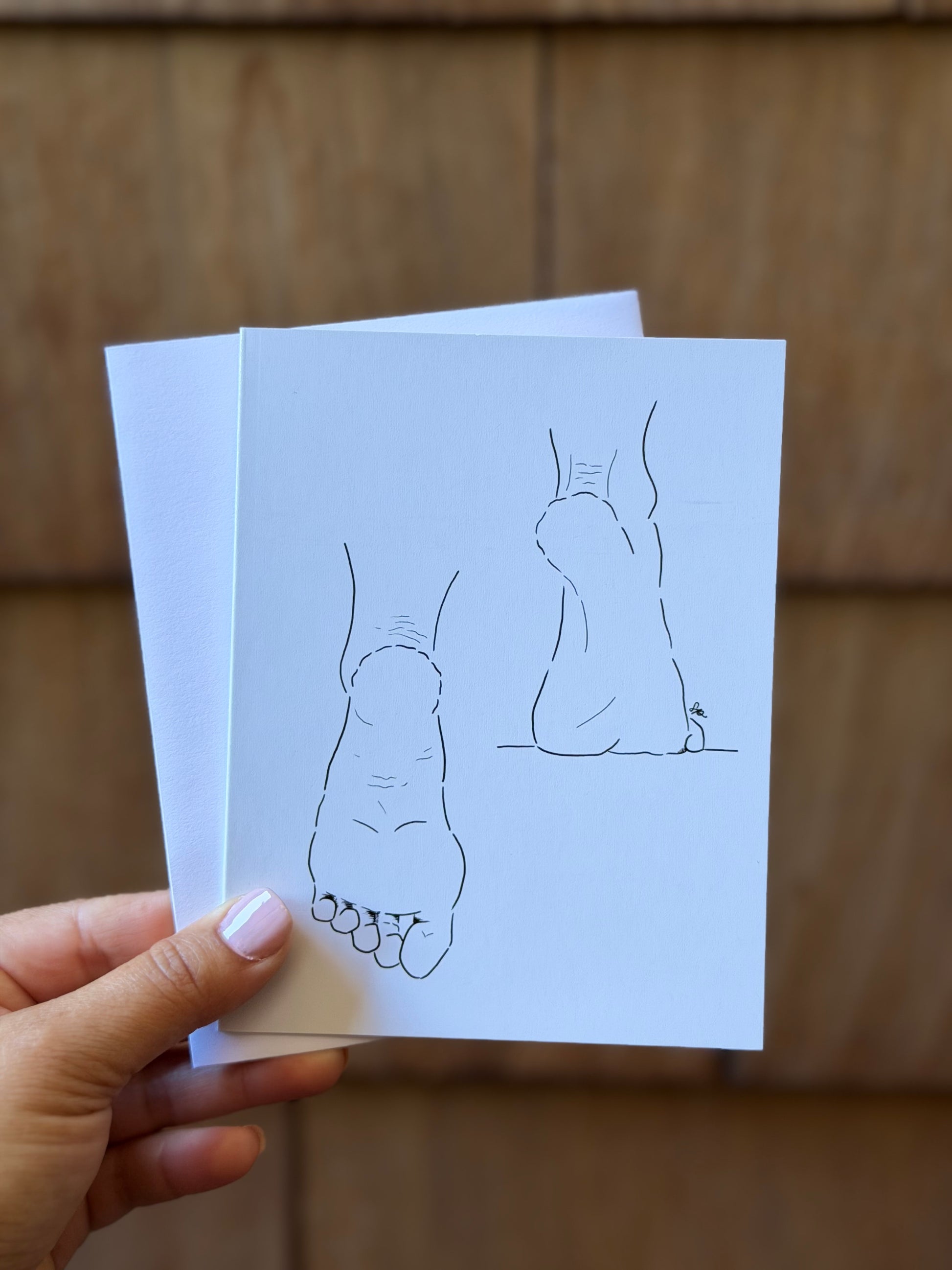 A hand with light pink nail polish holds a stack of white art prints featuring minimalist black line drawings of feet, set against a warm, blurred wooden background.