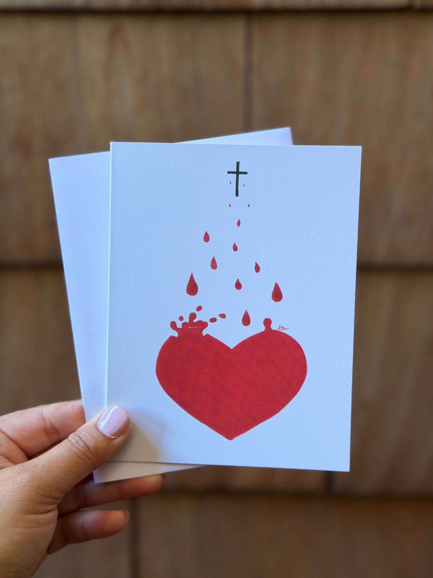 A hand holds a white greeting card featuring a red broken heart with red blood droplets and a small black cross above it, against a blurred brown wooden background.