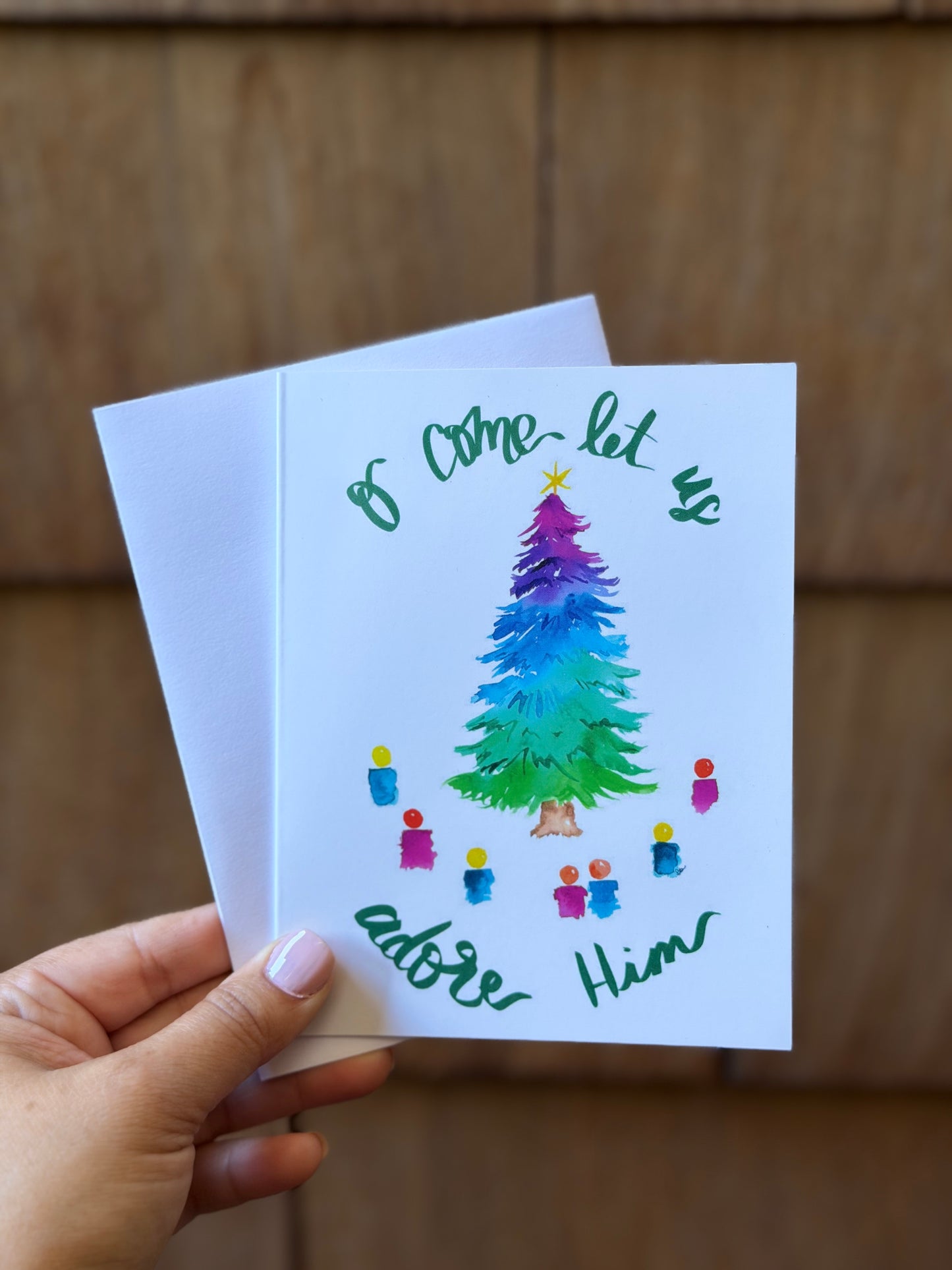 A hand holds a white greeting card with a colorful watercolor Christmas tree and green cursive text reading 'Come let us adore Him' against a brown wood-paneled background.