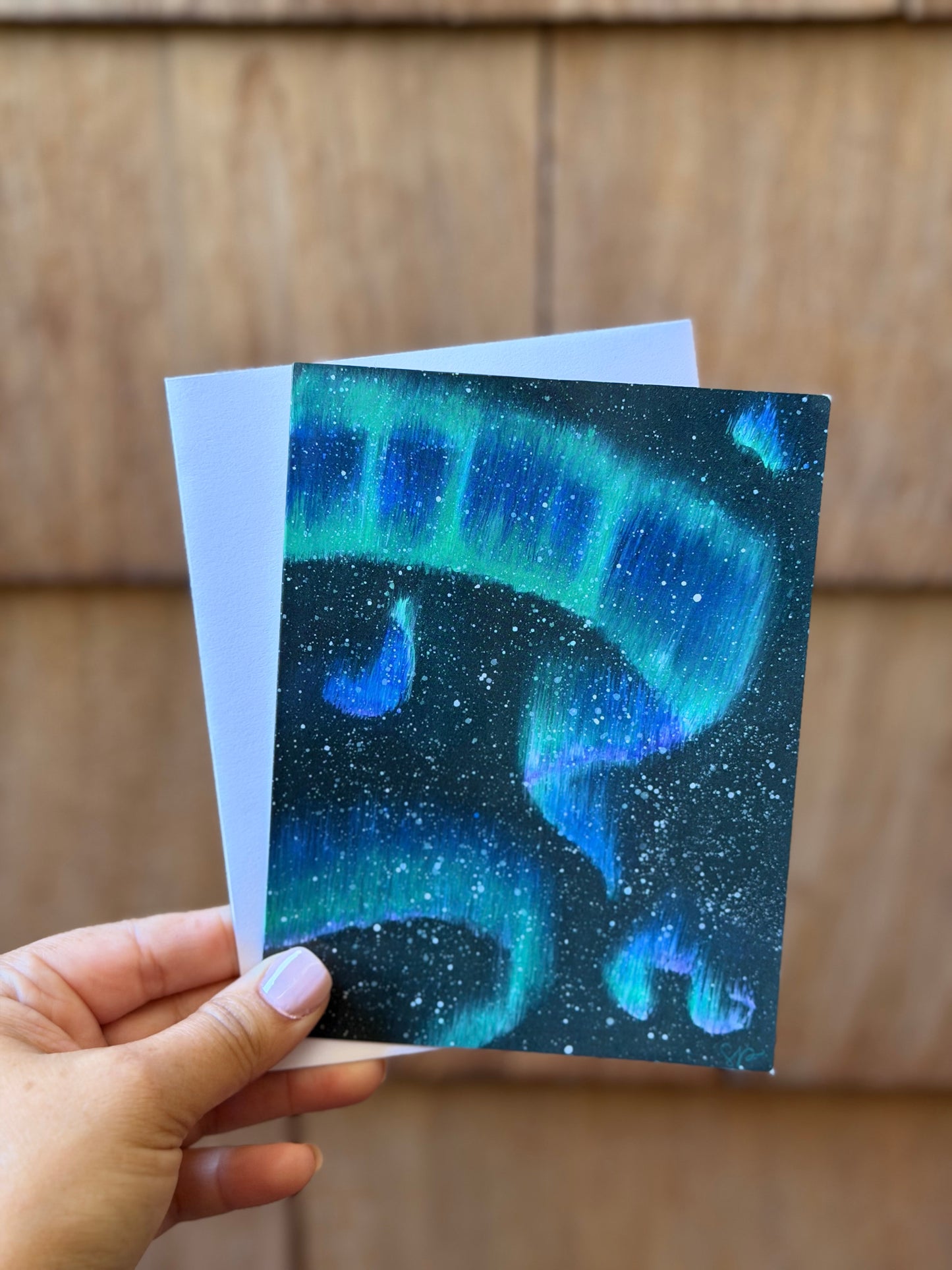 A hand holds a small card featuring a cosmic aurora design in shades of blue, green, and black, with a white backing card visible behind it, set against a wooden background.