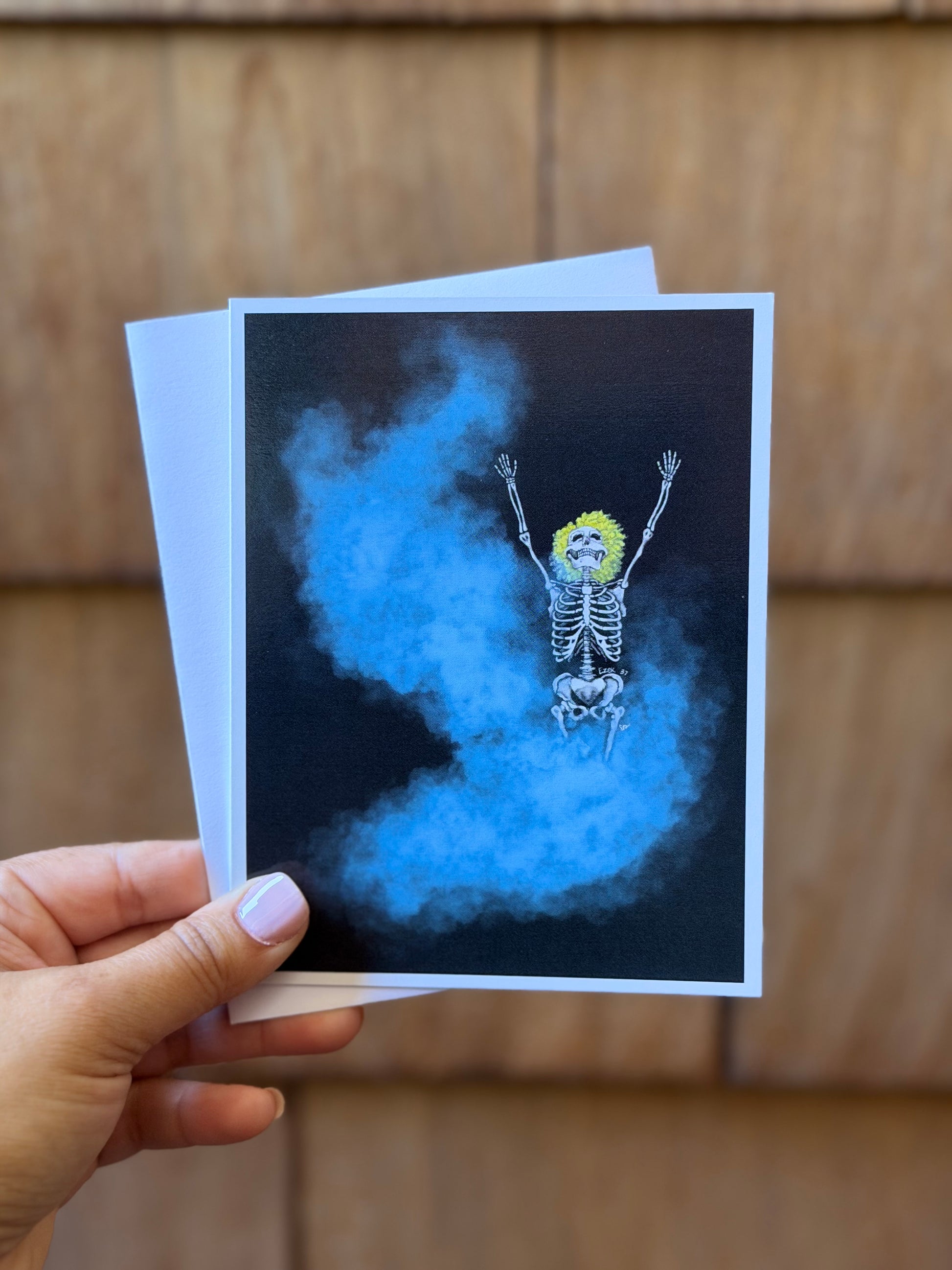 A hand holds a printed art card featuring a skeleton with a bright yellow wig floating in blue smoke against a dark background, with a wooden wall behind it.