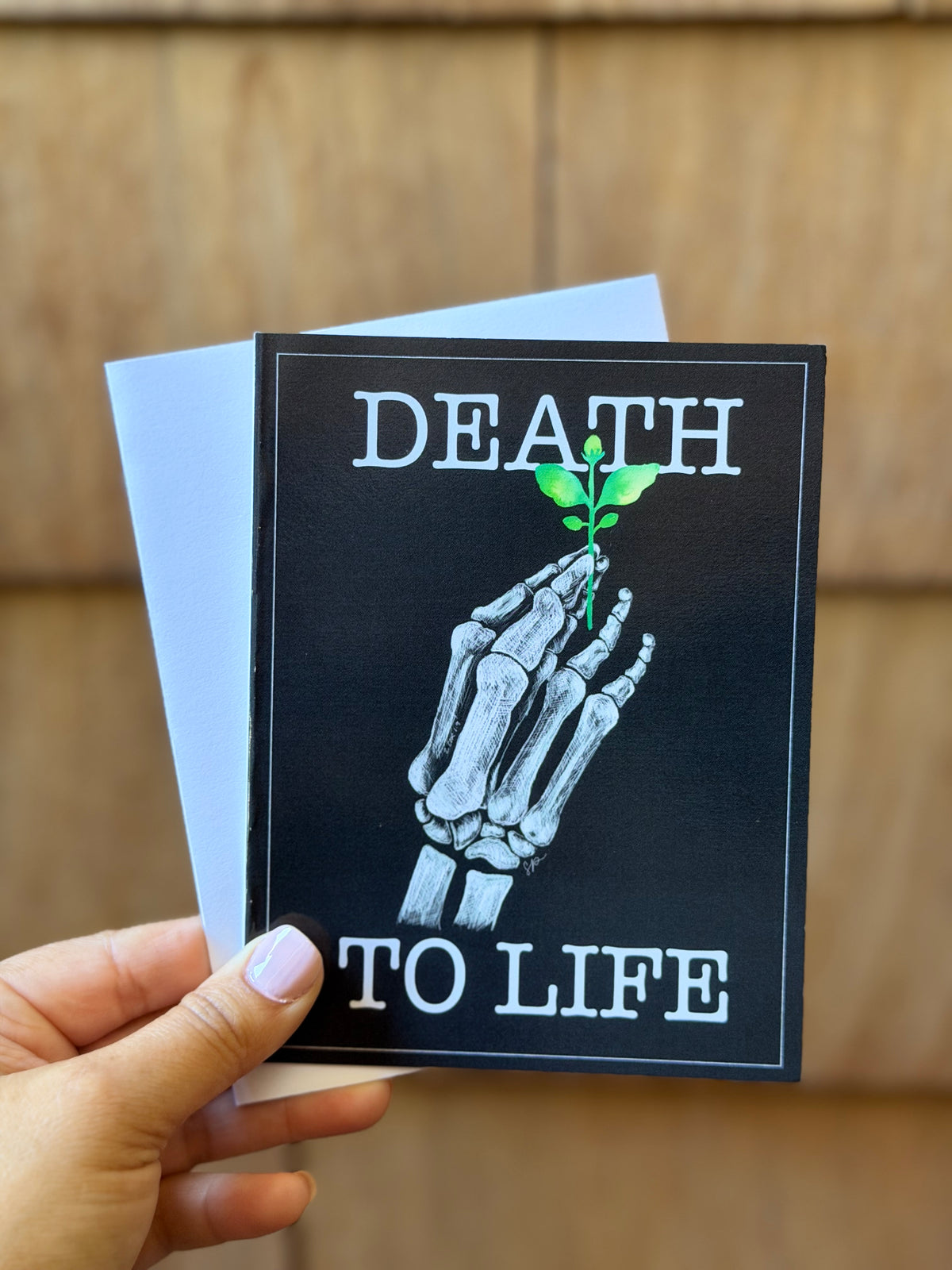 A hand holds a square black greeting card with the phrase 'DEATH TO LIFE' in white text and an illustration of a skeletal hand holding a small green plant. A white envelope is partially visible behind the card, against a blurred wood-paneled background.