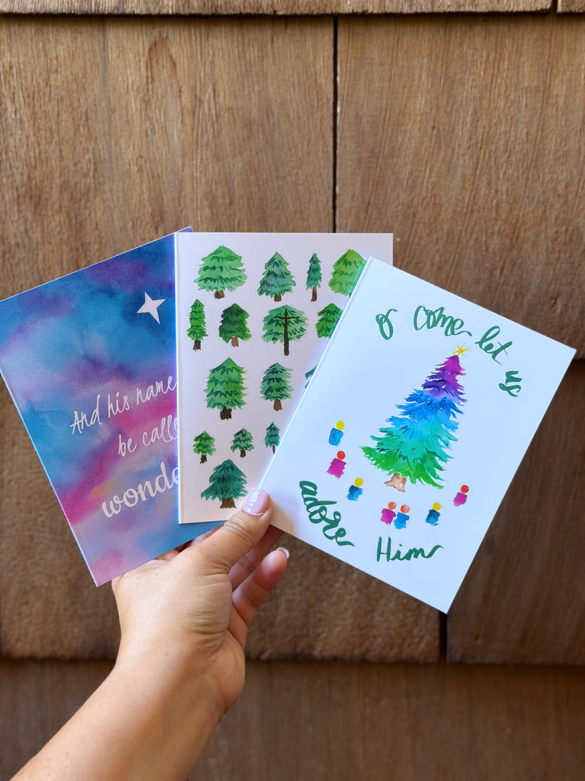 A hand holds three Christmas greeting cards against a wooden background. One card has a colorful watercolor sky design with white script text, another has green illustrated trees on a white background, and the front card shows a colorful watercolor Christmas tree with green script text.