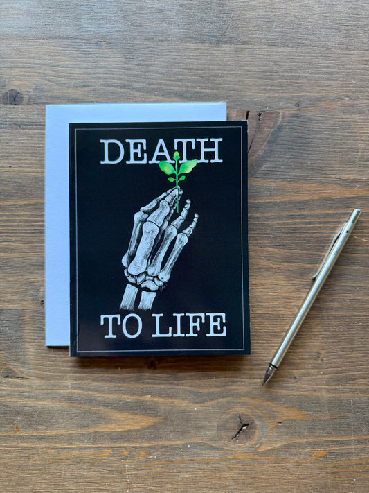 A square black greeting card with a white skeleton hand holding a small green plant, with the words 'DEATH TO LIFE' in white, placed on a wooden tabletop next to a white envelope and a silver pen.