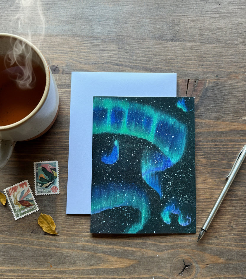 A square card featuring a vivid aurora borealis design in green and blue over a dark starry sky, displayed on a wooden tabletop next to a light blue envelope and a silver pen.
