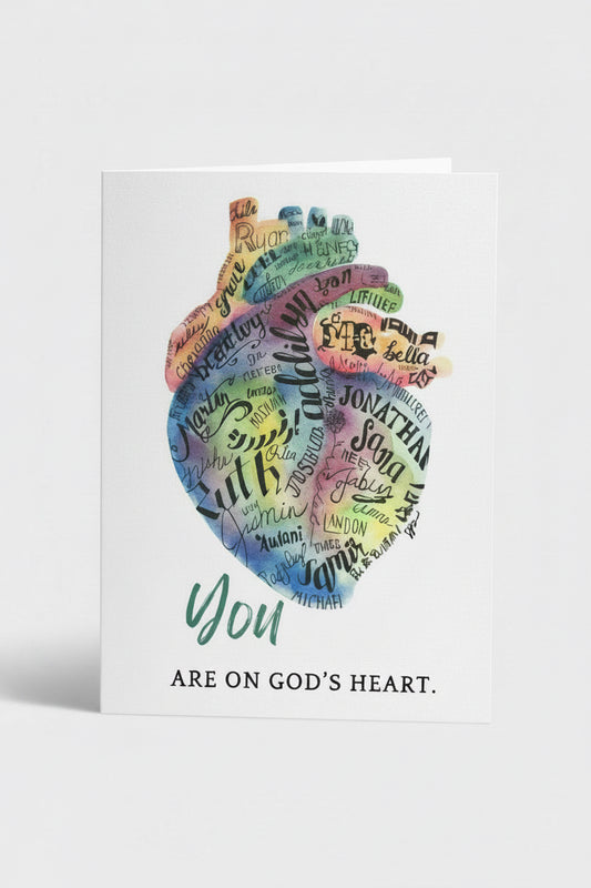 A white greeting card featuring a colorful watercolor heart made of handwritten Bible verses, with the words 'you ARE ON GOD'S HEART.' printed below, shown on a light neutral background.