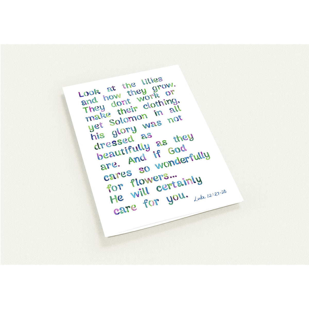 A white rectangular art card featuring a Bible verse from Luke 12:27-36 in multicolored lettering, photographed at an angle on a light off-white background.