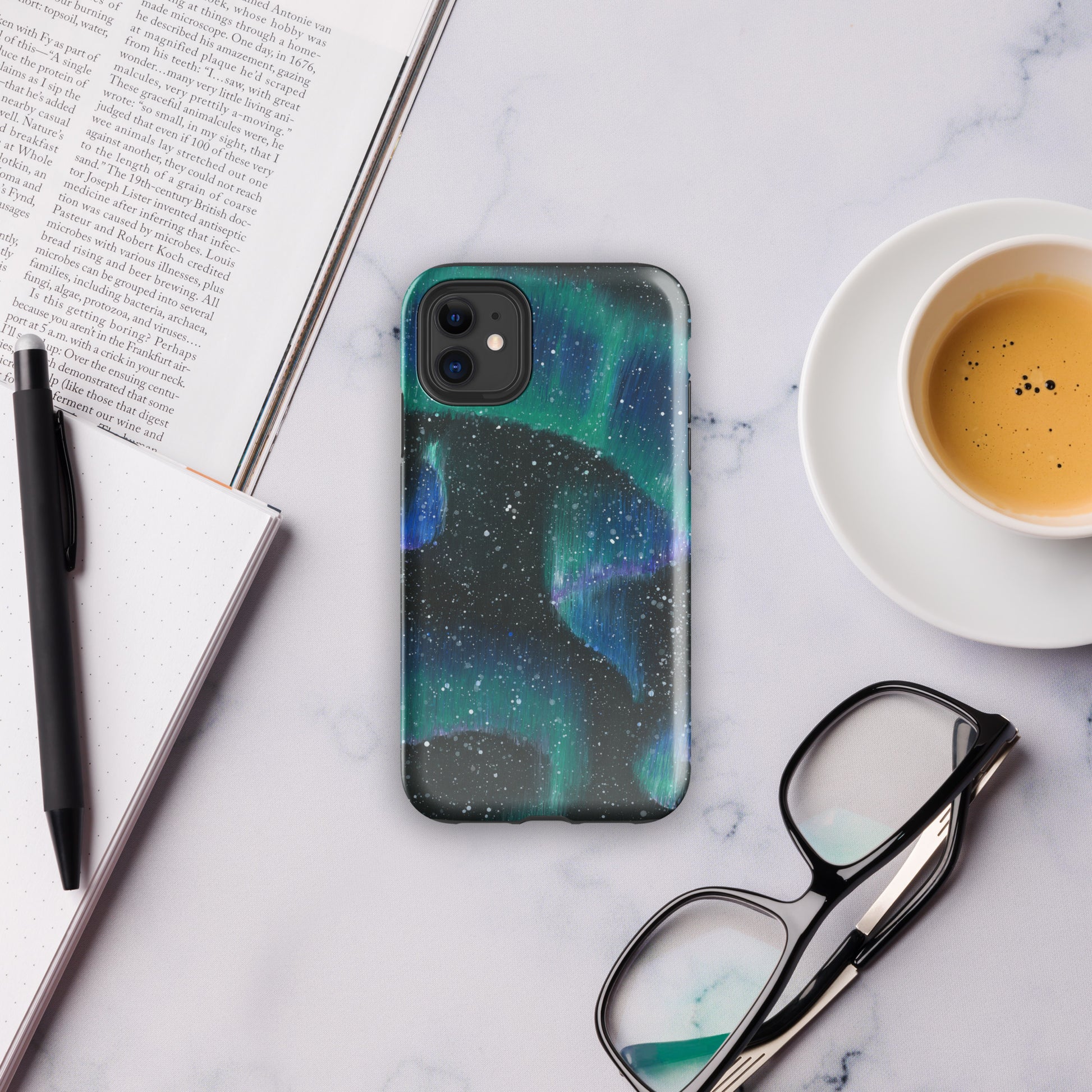 Mockup of phone case with northern lights design resting on a marble counter with a coffee, glasses, book, and notebook and pen